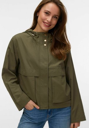 Uśmiechnięta kobieta z długimi brązowymi włosami, ubrana w oliwkowozieloną kurtkę z kapturem i niebieskie dżinsy, z ręką w kieszeni, na białym tle.