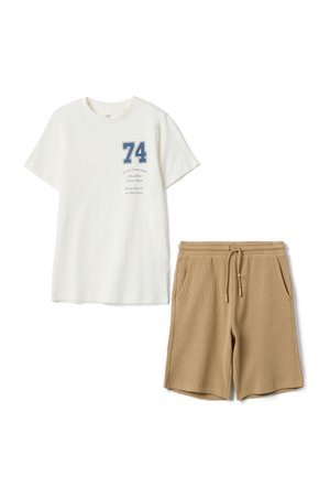 Weißes T-Shirt mit kurzen Ärmeln, blauem "74"-Aufdruck und kursivem Text, kombiniert mit hellbraunen Shorts mit Kordelzug und Fronttaschen.