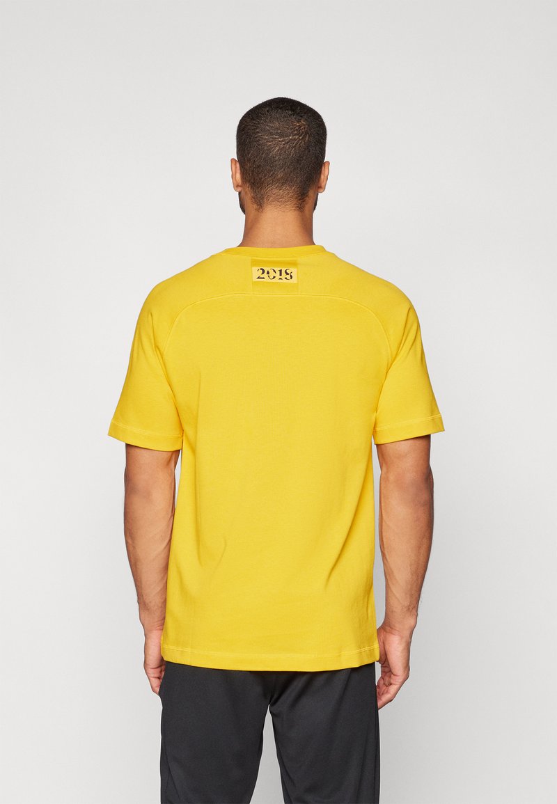 Uomo con capelli corti che indossa una camicia a maniche corte gialla brillante con il testo "2018" sulla parte superiore della schiena, abbinata a pantaloni neri.