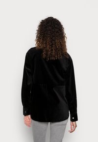 Camicia in corduroy nero con maniche lunghe, dotata di spallina posteriore e polsini con bottone, abbinata a pantaloni grigi chiari. Tessuto liscio, design minimal.
