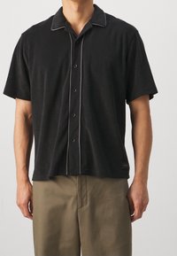 Lyhythihainen musta polo paita, jossa on pehmeä tekstuuri, nappisulku, vaaleanharmaa kontrastitikkaukset kauluksessa ja nappilistassa, yhdistettynä beigeihin housuihin.