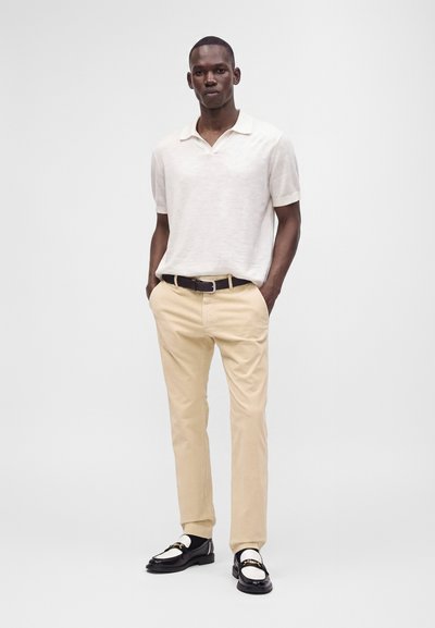 Homme debout les mains dans les poches, portant un polo blanc à manches courtes, un pantalon beige, une ceinture noire et des mocassins noirs et blancs.