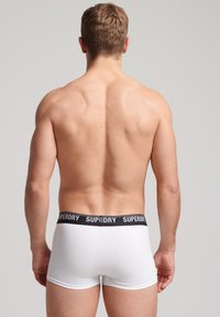 Bóxers blancos de ajuste ceñido con una cinturilla elástica negra etiquetada "SUPERDRY". Diseño simple, forma ajustada y textura suave. Sin patrones ni acentos.