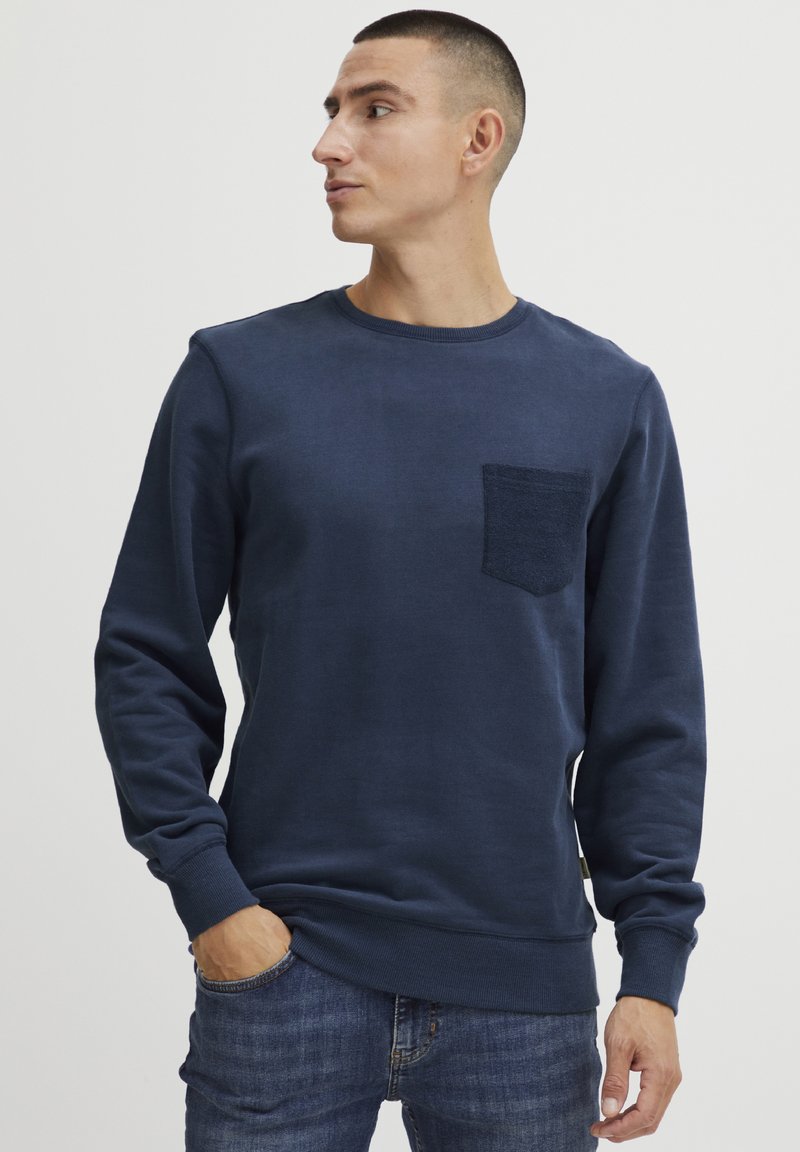 Felpa blu navy in cotone, con taschino a patch testurizzato, scollo rotondo e polsini e orlo a coste. Il modello la indossa con dei jeans.