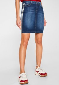 Falda de mezclilla en azul oscuro con franjas laterales blancas, con un diseño ajustado y un estilo clásico de cinco bolsillos, combinada con zapatillas blancas y rojas.