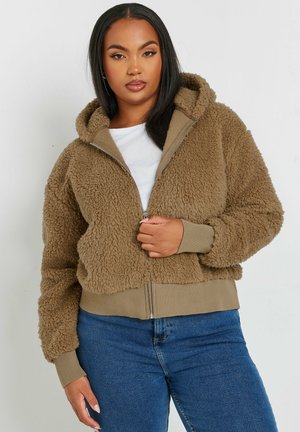 Fuzzy beige zip-up hoodie met een capuchon en ribgebreide manchetten, met een pluche textuur en een voorzak, gedragen over een wit T-shirt.