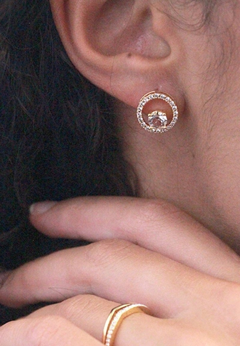 Orecchini circolari d'oro con una pietra centrale, circondata da piccoli cristalli trasparenti. Indossati nel lobo dell'orecchio, con una texture della pelle morbida visibile.