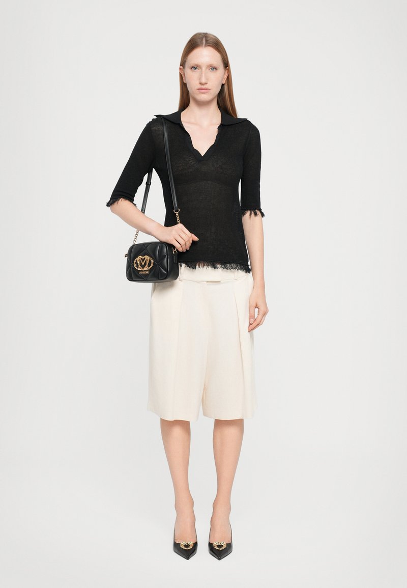 Top preto de malha com decote em V e mangas curtas, calções plissados em creme, mala preta com logótipo dourado, e sapatos de salto agulha pretos.