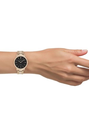 Montre en acier inoxydable doré avec un cadran noir rond, des index horaires minimalistes et un bracelet maillé, présentée sur une main.