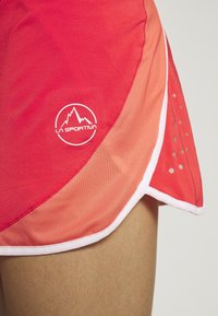 Nahaufnahme von roten und orangefarbenen La Sportiva-Sportshorts mit weißem Rand und reflektierenden Punkten an der Seite, getragen an einem bloßen Bein.