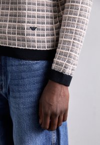 Pulover tricotat în nuanțe de bej și negru, cu un model în carouri. Prezintă mansete negre contrastante și un logo mic pe față. Asortat cu blugi albaștri.