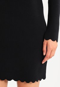 Black knitted dress with a ribbed texture, featuring a scalloped hem and long sleeves. Hand positioned on the dress.