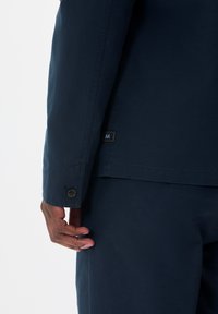 Jachetă bleu marin cu manșetă cu nasture, având o textură subtilă și o etichetă mică de marcă aproape de tiv.