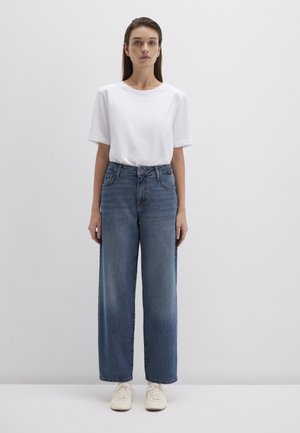 Weißes T-Shirt mit kurzen Ärmeln, kombiniert mit hoch taillierten, weit geschnittenen blauen Jeans und cremefarbenen Sneakers, steht vor einem schlichten weißen Hintergrund.