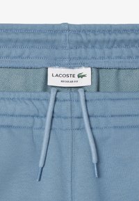 Pantalones de chándal azul claro con cinturilla elástica, que presentan una etiqueta blanca que dice "LACOSTE REGULAR FIT" y cordones para ajuste.