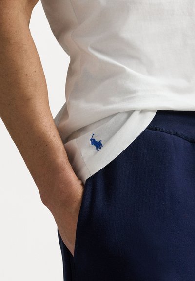 T-shirt en coton blanc avec un logo bleu sur l'ourlet, associé à un pantalon de sport bleu marine. Main visible dans la poche. Texture lisse.