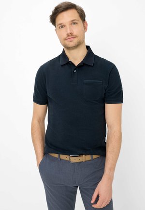 Homme aux cheveux bruns courts portant un polo bleu marine foncé et un pantalon gris avec une ceinture marron clair, debout les mains dans les poches.
