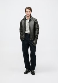 Czarna skórzana kurtka bombowa z zamkiem błyskawicznym, zestawiona z jasną koszulą w kratę, ciemnymi spodniami i czarnymi rękawiczkami. Całość uzupełniają ciemne, eleganckie buty.