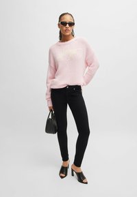 Maglione rosa chiaro a maglia con scollatura rotonda, abbinato a jeans skinny neri. Sandali neri con tacco alto e una piccola borsa nera completano il look.