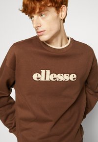 Sudadera marrón de tejido suave, con un logo bordado "ellesse" en color crema en el frente y un escote acanalado de contraste.