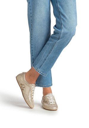 Zapatillas elegantes de color beige claro con parte superior de malla metálica, cordones blancos y detalles redondos; combinadas con vaqueros azules con puños doblados.