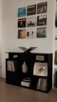 Unidade de armazenamento em madeira preta com vários compartimentos abertos, exibindo discos de vinil, um vaso e uma bolsa. Registros montados na parede acima apresentam várias capas de álbuns.