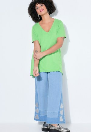 Mujer con pelo rizado que lleva una camiseta verde con cuello en V, vaqueros anchos azul claro con parches de corazones y zapatos de plataforma plateados, de pie sobre un fondo blanco.