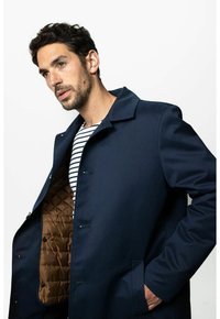 Marineblauer Mantel mit strukturiertem Kragen, offenem Vorderteil und braunem Steppfutter. Der Oberstoff wirkt glatt und ergänzt ein gestreiftes Innenhemd.