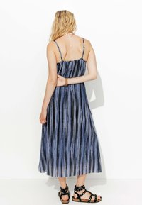 Robe maxi à fines bretelles, rayures verticales bleues et noires, et jupe fluide. Associée à des sandales à lanières noires.