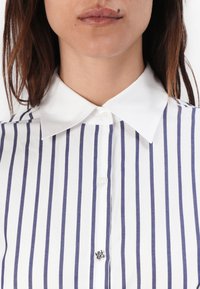 Chemise blanche avec des rayures verticales bleu marine, un col classique et une fermeture à boutons. Présente un petit logo imprimé près des boutons.