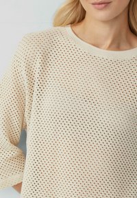 Top a maglia color crema con una trama a rete aperta, scollo rotondo e maniche corte. Il tessuto è leggero e traspirante.