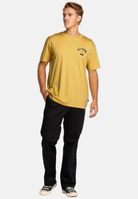 T-shirt gialla a maniche corte con logo nero e dettagli grafici, abbinata a pantaloni neri e sneaker. Design semplice, vestibilità rilassata.