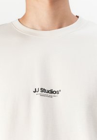 Lys beige bomulds t-shirt med rund halsudskæring, korte ærmer og et sort logo print af "JJ Studios" på forsiden.
