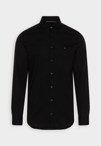 Camisa negra de manga larga con botones y cuello clásico. Presenta un logo sutil en el pecho, botones negros y una textura de tela suave.