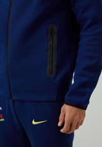 Mørkeblå atletisk jakke med sort lynlåslomme, fremstillet i struktureret stof og med lille gul Nike-logo på de matchende bukser.