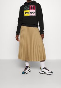 Černá mikina s kapucí a barevným potiskem, béžová skládací sukně, bílé a černé tenisky s barevnými akcenty a bílé kotníkové ponožky.