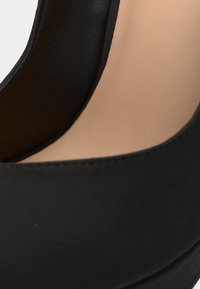 Chaussure noire à talon haut avec une texture lisse, un bout arrondi et une semelle intérieure rembourrée de couleur claire. Présente un design simple et élégant.