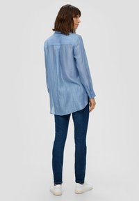 Helles blaues Langarmhemd aus glattem, strukturiertem Stoff, mit abgerundetem Saum und Knopfmanschetten, getragen mit dunkelblauen Jeans und weißen Schuhen.
