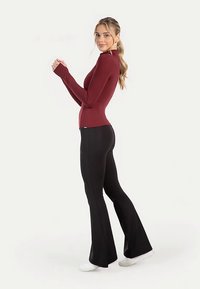 Bordeauxrotes, langärmeliges Oberteil kombiniert mit schwarzen, ausgestellten Fitnesshosen. Oberteil: Figurbetontes Design, weiche Textur. Hose: Dehnbarer Stoff, glatte Oberfläche, dazu weiße Sneakers.