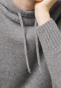 Szary sweter dziany z wysokim kołnierzem, długimi prążkowanymi rękawami i sznurkami ściągającymi. Materiał wydaje się miękki i przytulny.