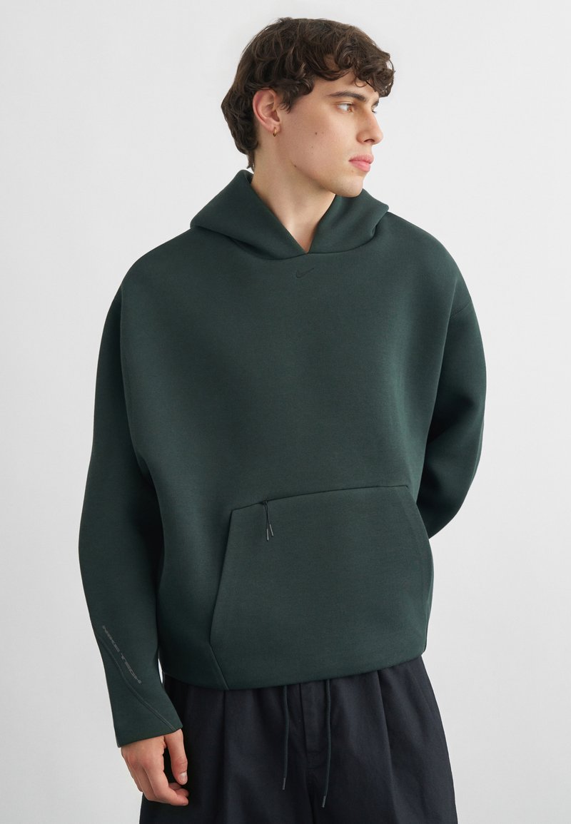 Jeune homme aux cheveux bouclés portant un sweat à capuche vert foncé oversize et un pantalon noir, regardant vers la droite sur un fond blanc uni.