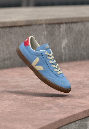 Baskets en daim bleu avec des lacets crème et un logo, un passant de talon rouge, et une semelle marron flottant au-dessus d'un sol en pavé de pierre.