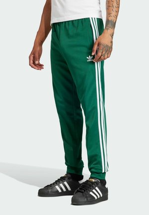 Homme portant un pantalon de survêtement Adidas vert avec des bandes blanches et des baskets Adidas noires à bandes blanches, debout sur un fond clair.