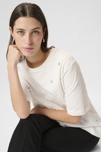 Tricou din bumbac crem, cu detalii decorative aurii rotunde, mâneci scurte și o croială relaxată, asortat cu pantaloni negri. Textură simplă și netedă.