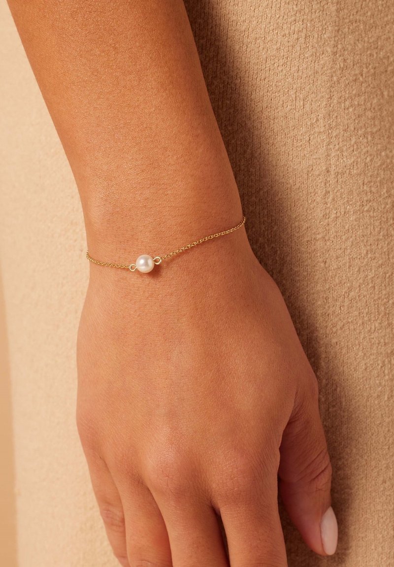 Gold bracelet featuring a small white pearl at the center, resting on a wrist against a textured beige background.