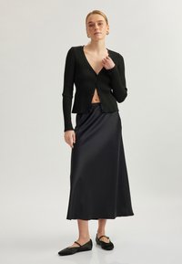 Schwarze gerippte Strickjacke mit einem tiefen V-Ausschnitt, kombiniert mit einem schwarzen Satin-Maxirock. Schwarze Ballerinas runden das Outfit ab. Glatte Textur und figurnahe Passform.
