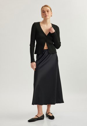 Cárdigan negro de canalé con un profundo escote en V, combinado con una falda maxi de satén negra. Unos zapatos planos negros completan el conjunto. Textura suave y diseño ajustado.