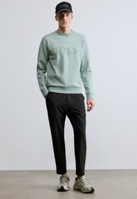 Világoszöld pulóver dombornyomott "BOSS" logóval, fekete nadrággal és fehér sportcipővel párosítva. A modell fekete sapkát visel logóval.