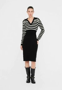 Črno-bela črtasta obleka pulover z V-izrezom, dolgimi rokavi in prilegajočo silhueto. Ponaša se z detajli logotipa in črnimi škornji do kolen.