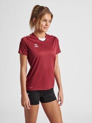 Kvinde iført vinrød sportsskjorte og sorte shorts, stående med afslappede hænder, kiggende let mod højre mod en ensfarvet baggrund.